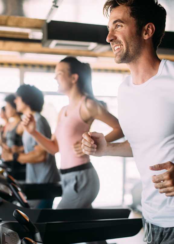 A man in a white t-shirt is running on a treadmill in a gym with other people exercising around him.