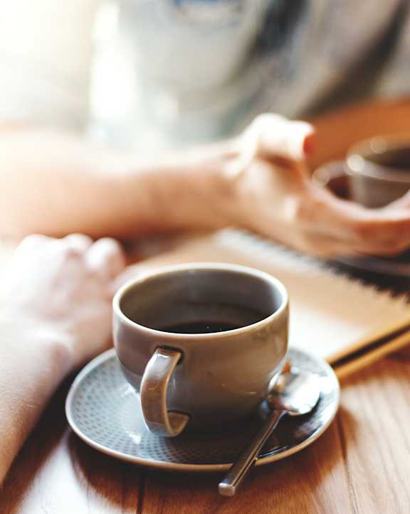 A person is holding a cup of coffee in one hand while the other hand is on a table with a notebook and a cup of coffee.