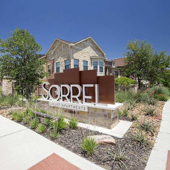 The entrance at Sorrel Luxury Apartments sign, surrounded by bushes and trees, with a building in the background. 