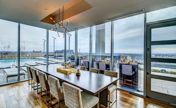 A modern dining room with a long wooden table and chairs, a chandelier, and a view of the outdoors through large windows.