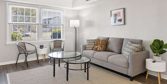 A living room with a grey couch, a glass table, and a window with a view of the street.