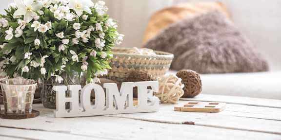 A white wooden table with a white flower pot, a white HOME sign, and a brown knitted pillow.