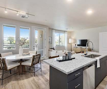 The spacious living room with a white couch, a balcony, large windows, white plush chairs, coffee table, decorations, a TV, and view of the dining room with a white dining room and white table and the kitchen with marble counters and stainless-steel appliances at Reflect at Dobson Ranch Apartment Homes. 