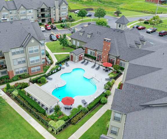 The arial view of the large, turquoise outdoor pool with pool chairs, tables, and chairs surrounded by green space with the apartments in the background at Villas at Preston Lakes Apartment Homes. 