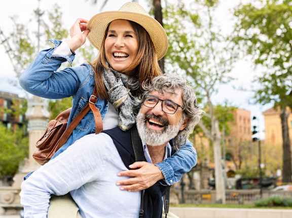 A woman in a hat is giving a piggyback ride to an older man in a city park.