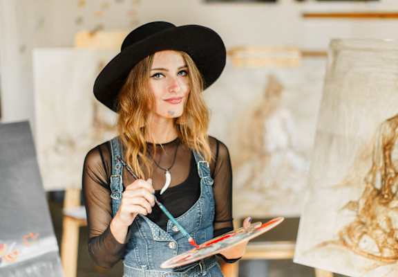 a woman in a hat holding a paintbrush and a canvas