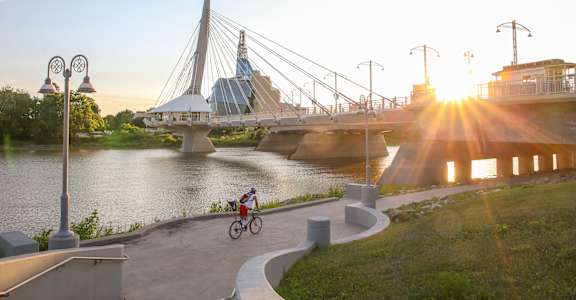 A cyclist rides along a path next to a river under a bridge.