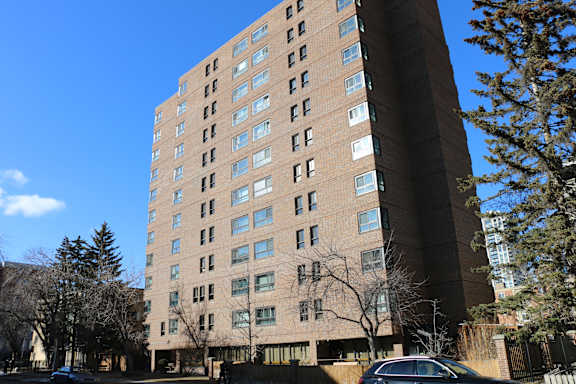 A tall building with many windows is surrounded by trees and a car is parked in front.