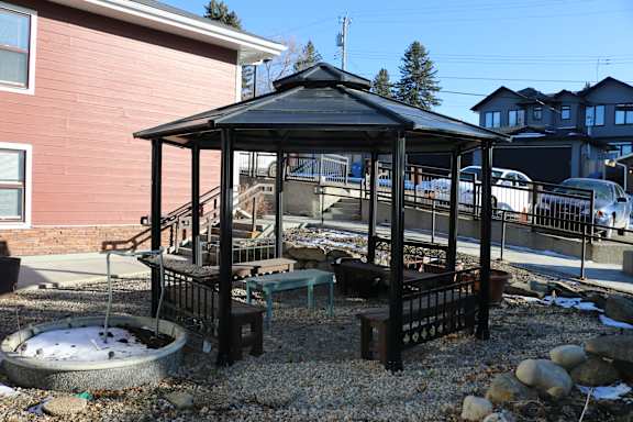 A gazebo with a black roof is surrounded by a gravel area with a small pond and a bench.