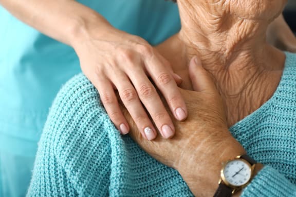 A close up image of an elderly woman holding someone else's hand on their shoulder