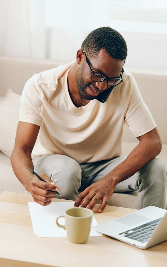 A man is writing on a piece of paper with a pen while sitting on a couch with a laptop and a cup of coffee on the table in front of him.
