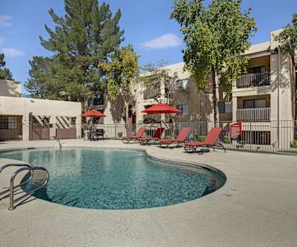 The turquoise outdoor pool with pool chairs, tables, and chairs with the apartments in the background at Paradise Falls Apartments. 