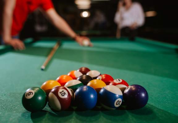 A pool table with a variety of balls and a person's arm holding a cue.