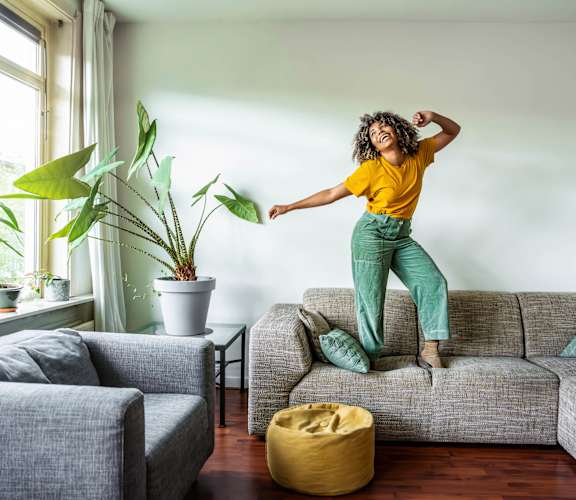A woman in a yellow shirt and green pants is jumping on a grey couch in a living room with a yellow pouf and potted plants.