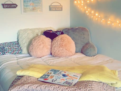 Gray, pink, and white fluffy pillows cover a full extra long bed that is 54 by 80 inches. There is an off-white blanket covering the bed and a yellow throw blanket at the end of the bed. Decorative wall signs are spread throughout the space.