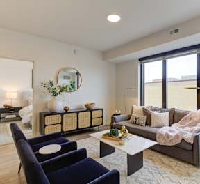 A living room with a grey couch, a blue chair, a white table, and a round mirror on the wall.