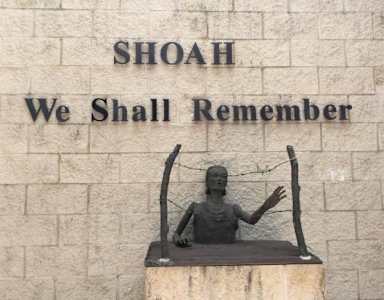 A statue of a person with outstretched arms is placed in front of a wall with the words "SHOAH We Shall Remember" engraved on it.