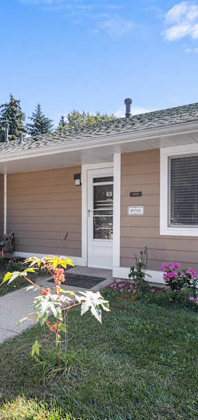A small house with a brown and white exterior is surrounded by greenery and flowers.