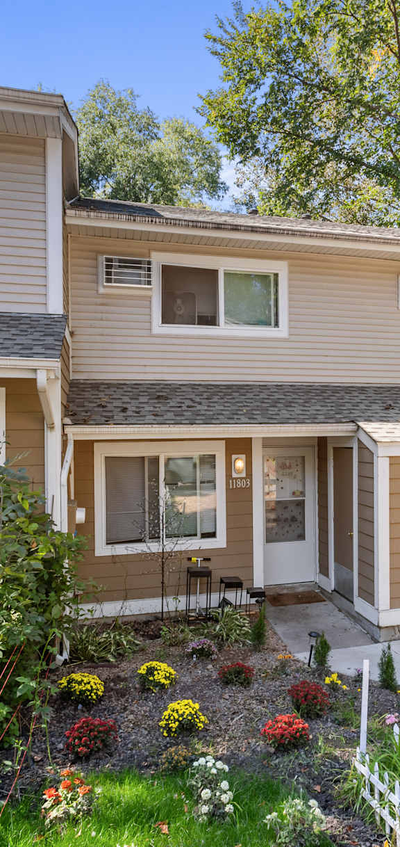 A house with a brown and beige exterior is surrounded by greenery and has a small garden in front.