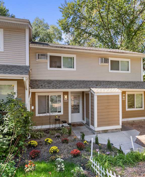 A house with a brown and beige exterior is surrounded by greenery and has a small garden in front.