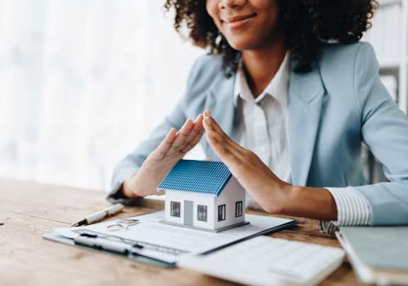 A woman in a blue blazer is sitting at a desk with a small model house, papers, and a pen.