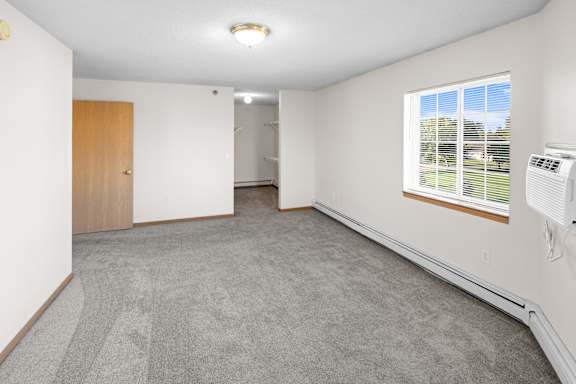 A room with a carpeted floor, a window with blinds, and a wall-mounted air conditioner unit.