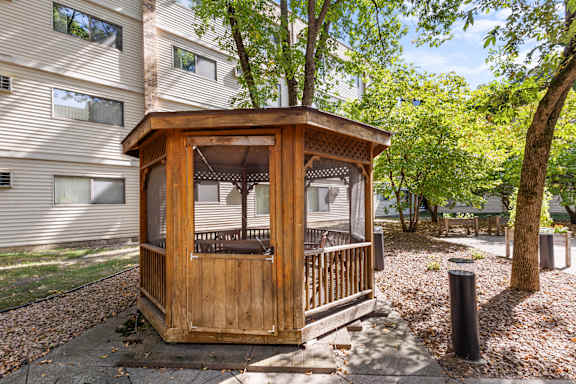 A wooden gazebo is situated in a grassy area in front of a building.