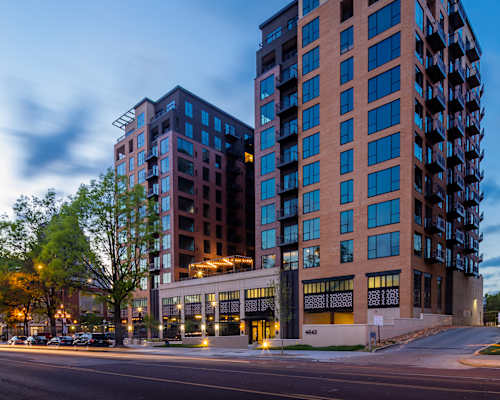 A modern multi-story building with a glass facade is illuminated from within, with a tree and a street in the foreground.