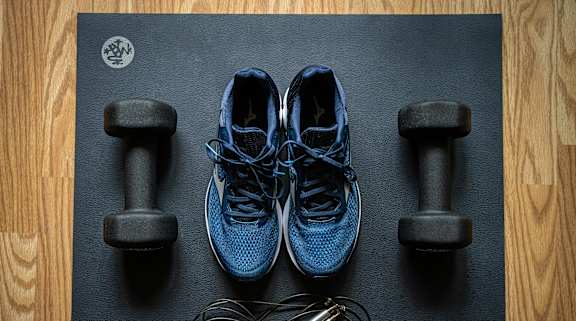 A pair of blue sneakers, two dumbbells, and a skipping rope are laid out on a black mat.