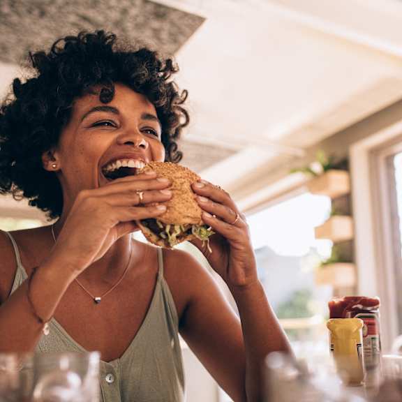 A woman with curly hair is laughing while eating a sandwich.