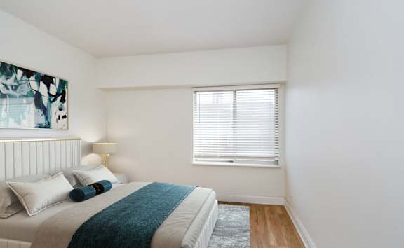 A bedroom with a bed, pillows, a blanket, a lamp, and a window with blinds.