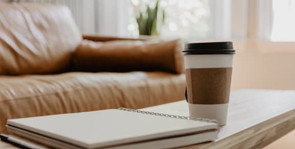 Tan leather couth with wooden coffee table in front. On the wooden coffee table is a pen, notepad, and to go coffee cup.