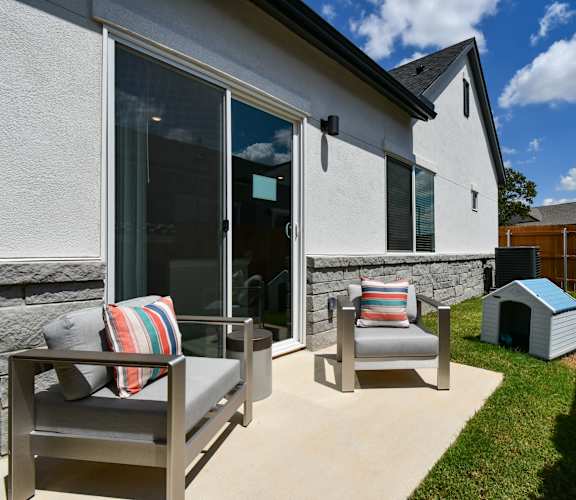 A patio with a grey couch and a striped pillow is in front of a house.