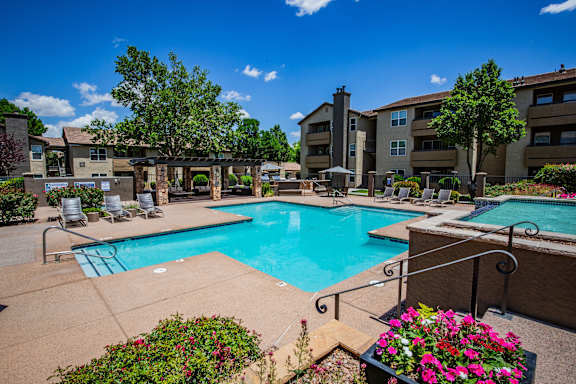 A pool surrounded by chairs and flowers with apartment buildings in the background.