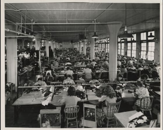 A black and white photo of a factory with workers seated at desks.