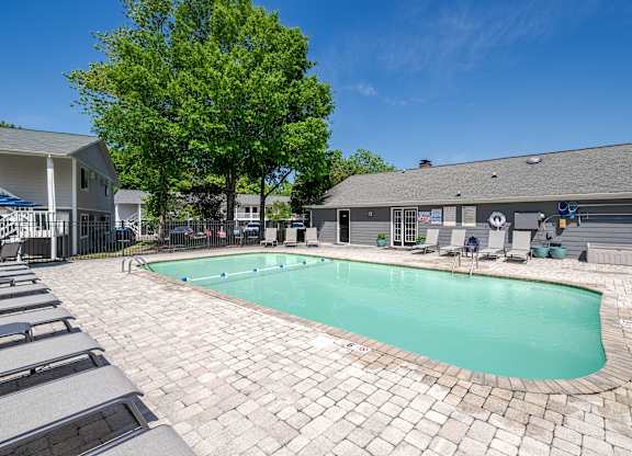 Outdoor swimming pool with crystal-clear water, surrounded by a paved deck with lounge chairs and shaded seating, set against a clubhouse and lush green trees under a bright blue sky