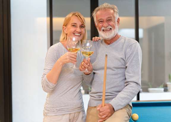A man and a woman are standing by a pool table, each holding a glass of wine.