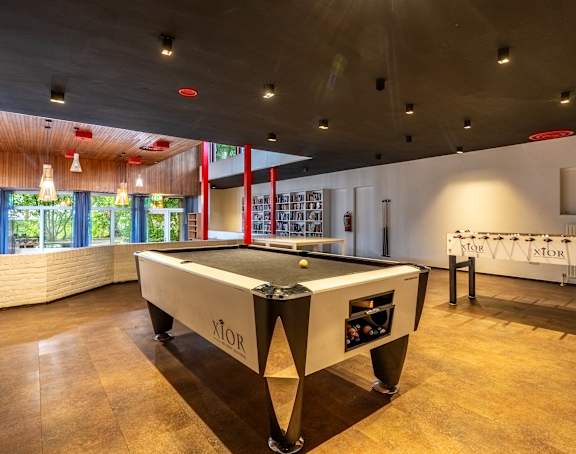 A pool table with the word XTOR on it is in the middle of a room with a wooden floor and a window with curtains.