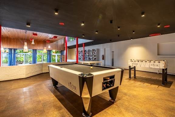 A pool table with the word XTOR on it is in the middle of a room with a wooden floor and a window with curtains.