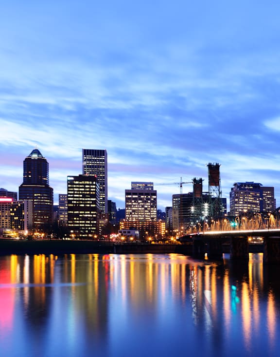 A city skyline at dusk with a bridge over a calm body of water.