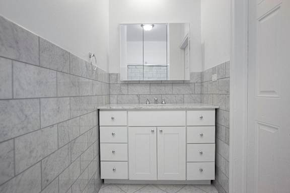 A white bathroom with a marble tile wall and a white vanity with a mirror above it.at Sutton Collection, New York New York