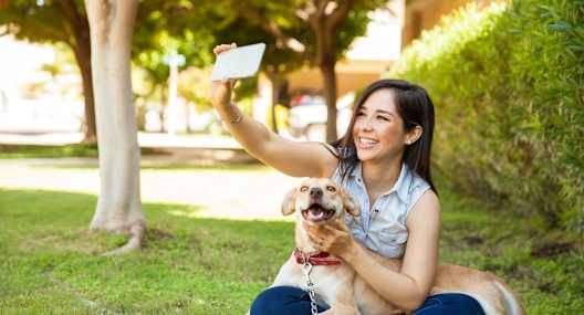 A woman is sitting on the grass with her dog, taking a selfie.
