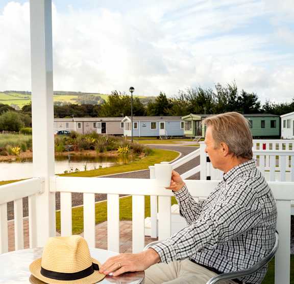 A man in a plaid shirt and hat sits on a porch with a cup in hand.