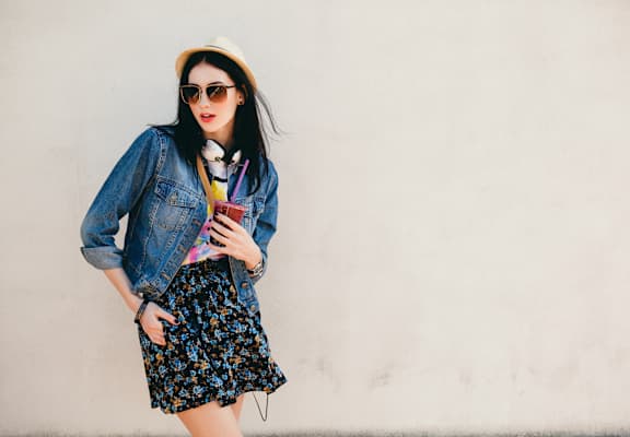 A woman in a floral skirt and denim jacket is holding a drink.