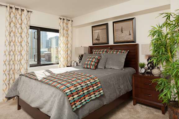 A bedroom with tan walls and carpet, and a bed with a grey bedspread and colorful throw