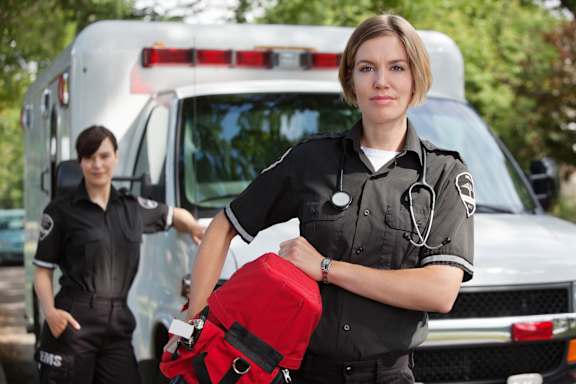 A woman in a black uniform is holding a red bag in front of an ambulance.
