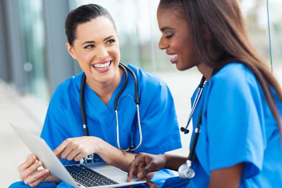 Two medical professionals in blue scrubs are looking at a laptop screen together.