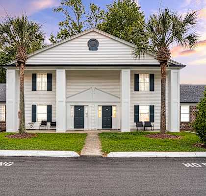 A white building with a black door and windows is surrounded by greenery.