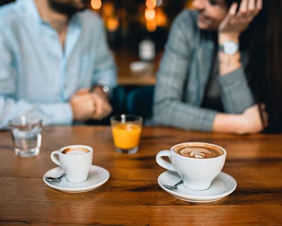 Two cups of coffee are on a table with a glass of water and a glass of orange juice in the background.