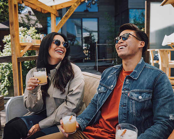 Two people are sitting on a couch, each holding a drink, and laughing together.